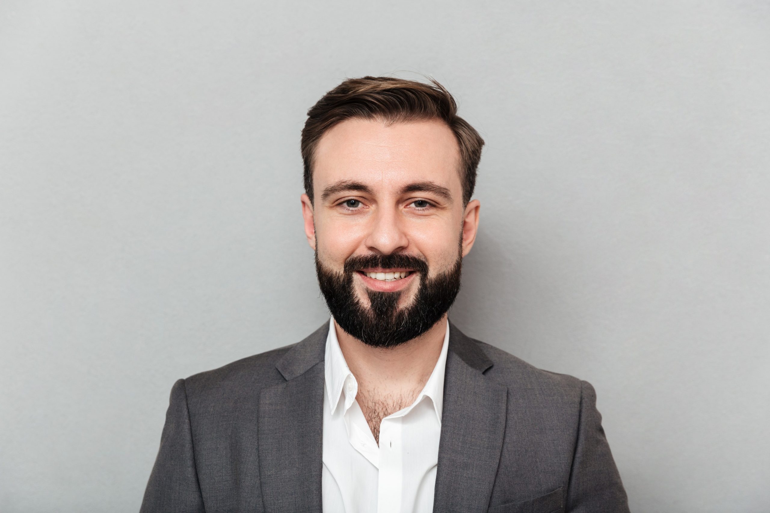 close-up-portrait-young-bearded-man-white-shirt-jacket-posing-camera-with-broad-smile-isolated-gray-1-1-scaled.jpg